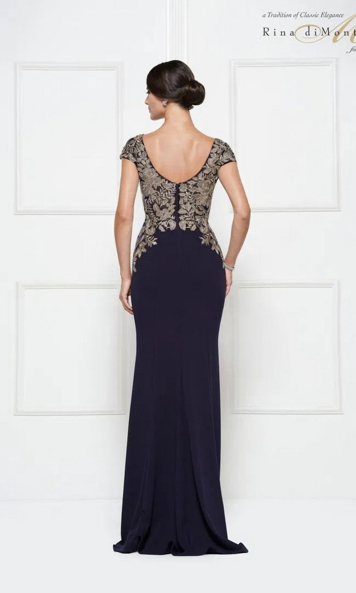 A woman faces away from the camera in the Rina Di Montella RD2652, a floor-length navy formal dress with gold embroidery on the upper back and cap sleeves.